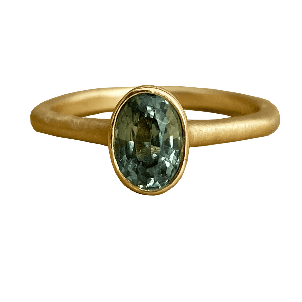 A teal-green-blue sapphire engagement ring with a unique bezel setting. A hidden diamond sparkles beneath the sapphire.