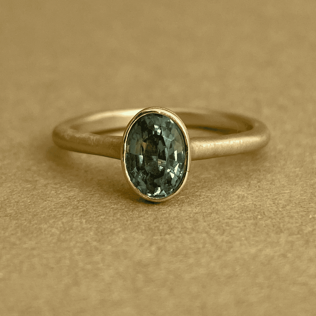 A teal-green-blue sapphire engagement ring with a unique bezel setting. A hidden diamond sparkles beneath the sapphire.