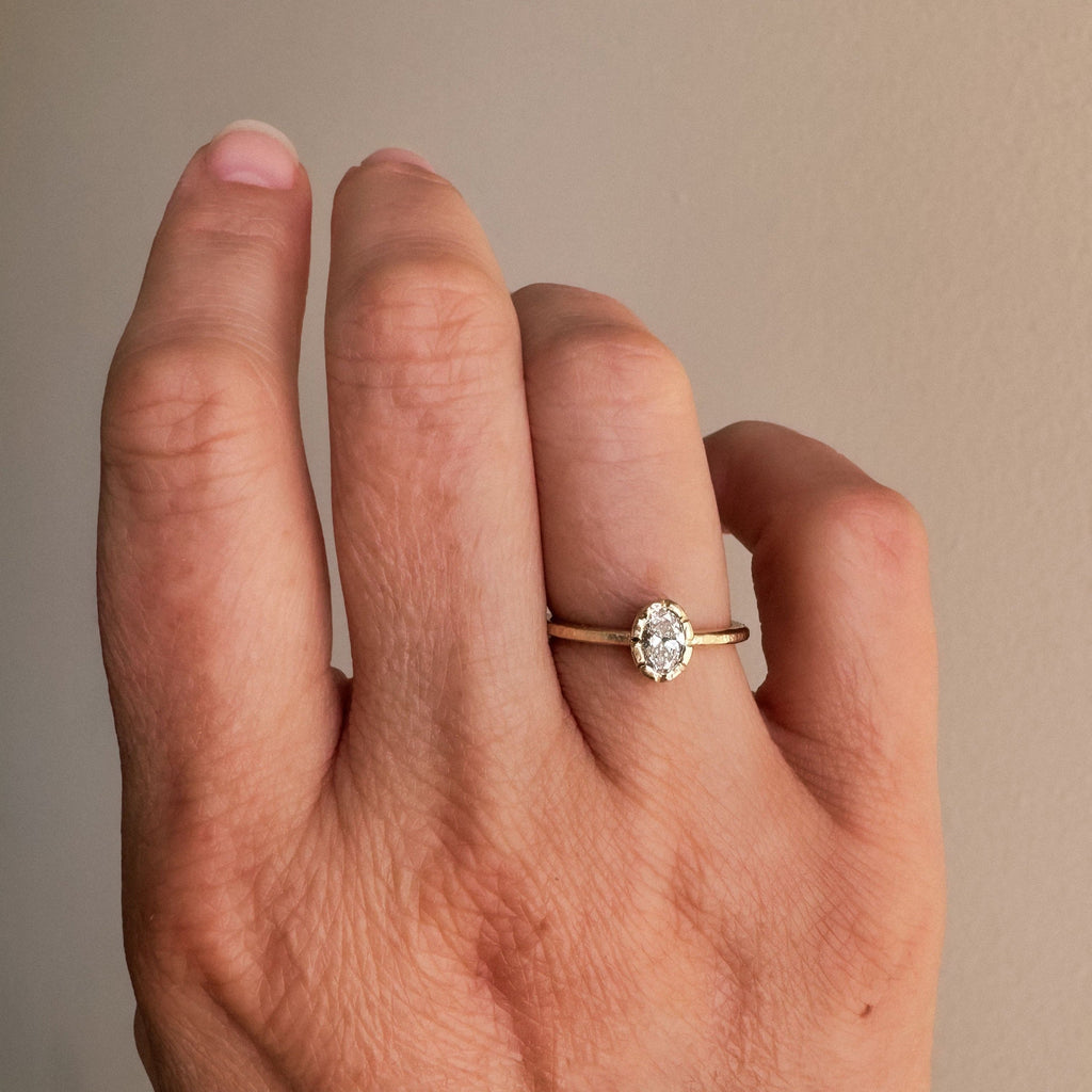 A close-up of a woman's hand wearing a delicate engagement ring with a unique bezel setting and an oval lab diamond. The ring is inspired by a wildflower and symbolizes the beauty and tenderness of nature's embrace.