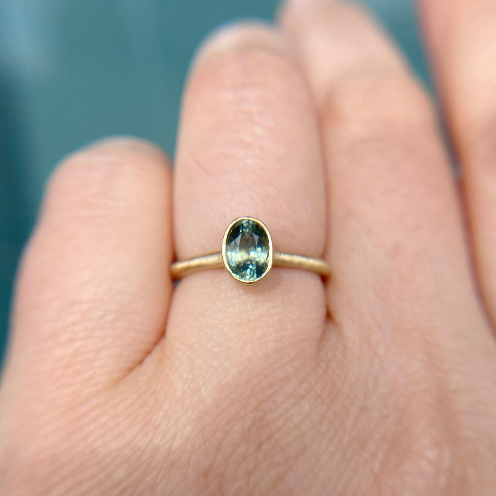 A teal-green-blue sapphire engagement ring with a unique bezel setting on a woman's finger. A hidden diamond sparkles beneath the sapphire.